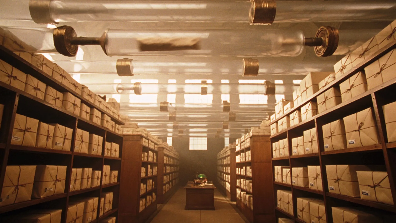 The long archive hall of the Dead Letter Office, shelves of parcels rising into dusty golden light beneath glass pneumatic tubes